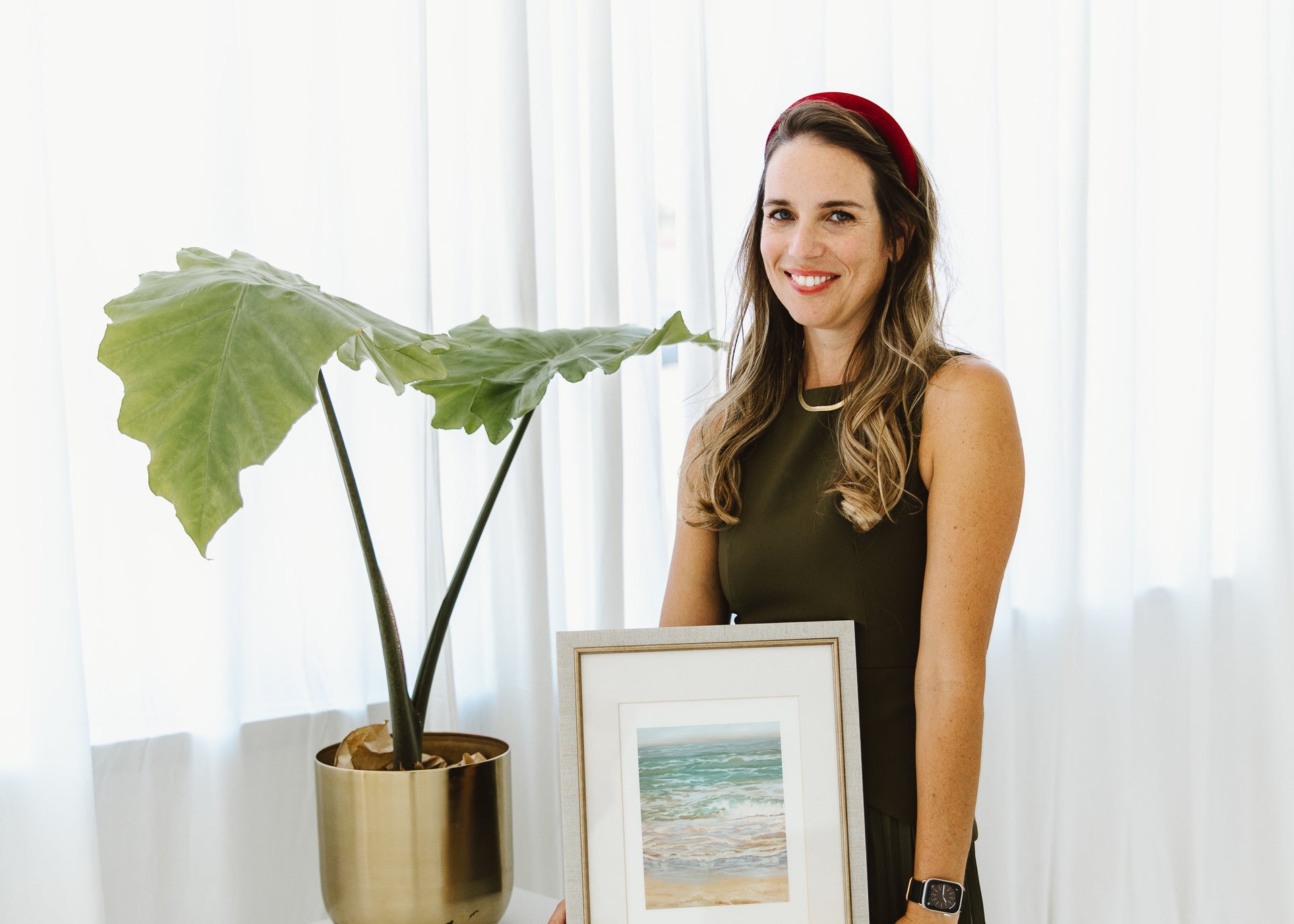 A woman in a black sleeveless dress and red headband stands beside a white table holding a framed beach landscape print. A potted green plant in a gold pot sits on the table against a bright white curtain background.