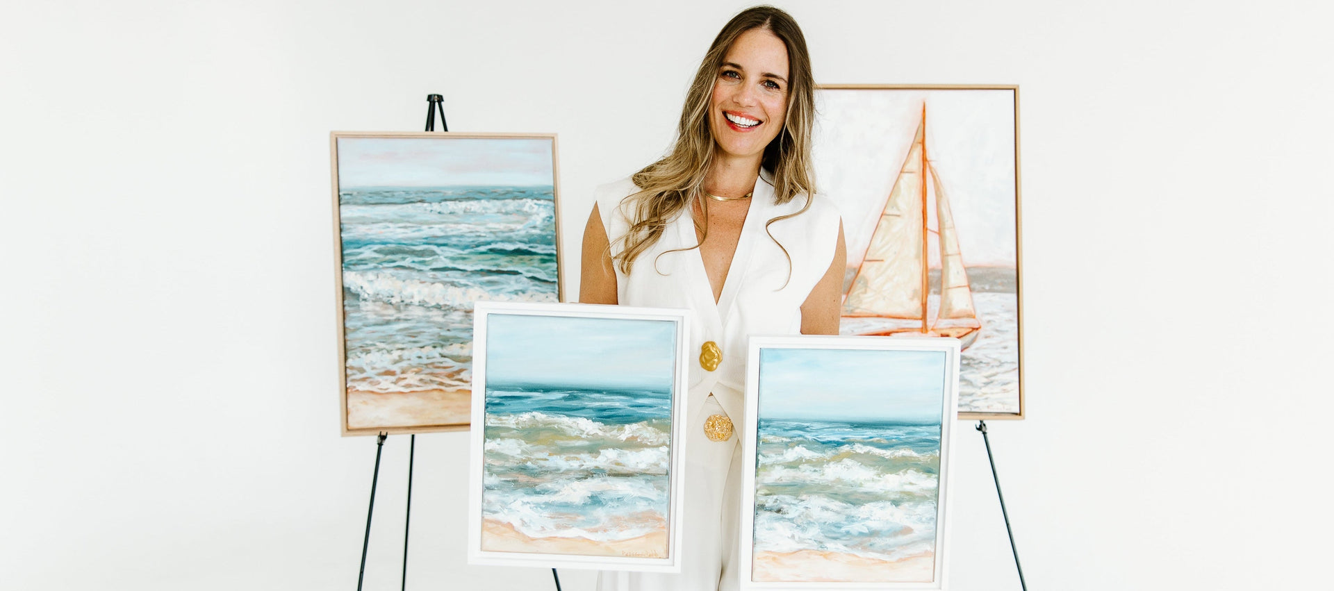 A woman in a white jumpsuit stands in a bright white studio holding and displaying several framed beach-themed paintings on easels.