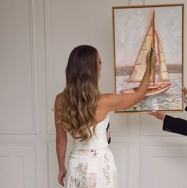 A woman stands with her back to the camera wearing a white strapless midi dress with a printed sailboat and architectural scene pattern. She is holding a framed painting of a sailboat next to a man's arm in a white paneled room.