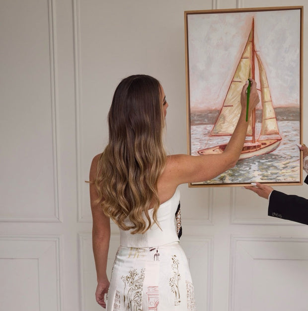A woman stands with her back to the camera wearing a white strapless midi dress with a printed sailboat and architectural scene pattern. She is holding a framed painting of a sailboat next to a man's arm in a white paneled room.